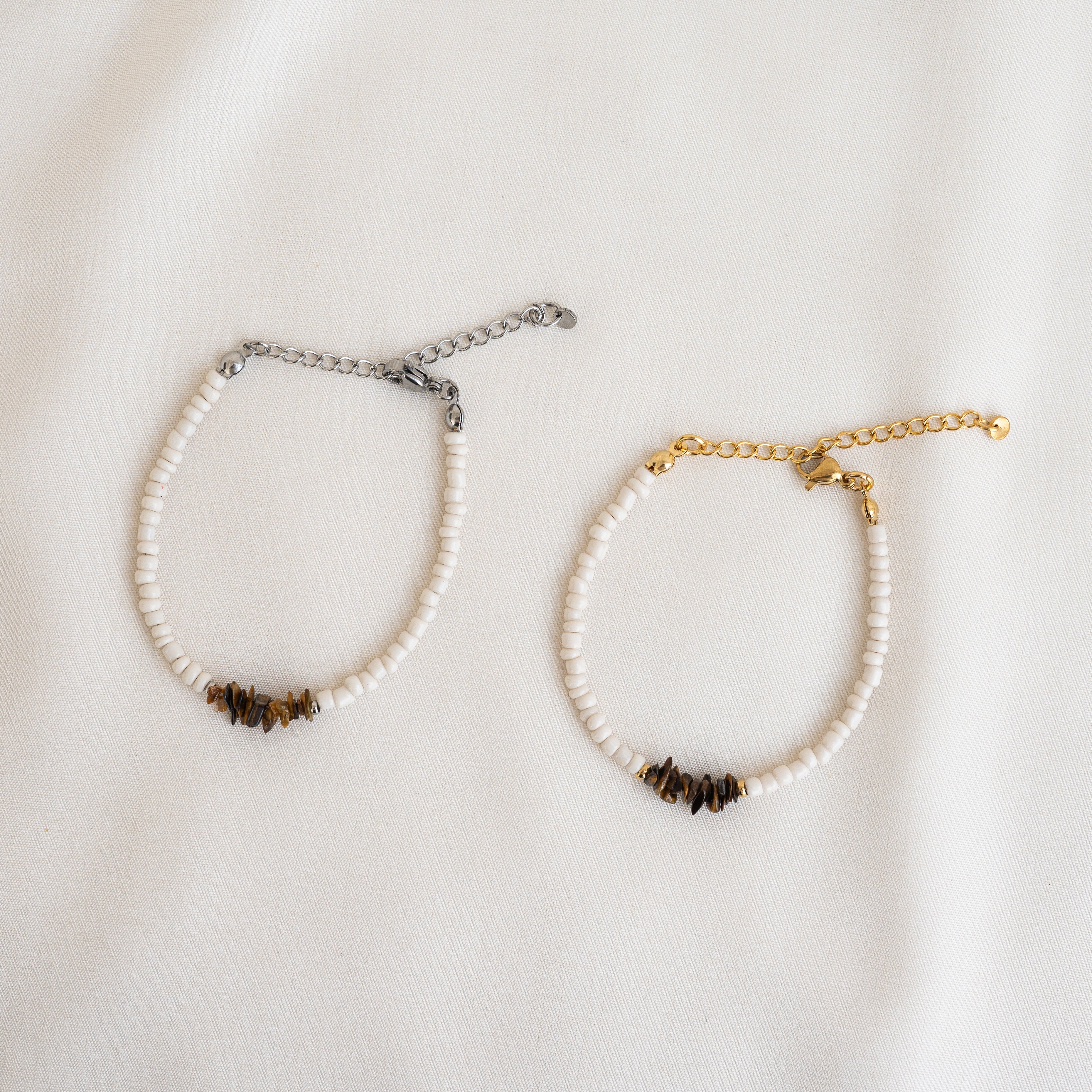 Zwei Perlenarmbänder mit weißen Perlen und kleinen dunklen Akzentperlen; eines hat einen silbernen Kettenverschluss, das andere einen goldenen Kettenverschluss, beide auf weißem Stoffgrund.