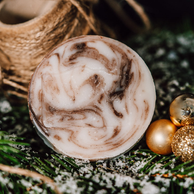 Runde Seife mit braunen Wirbeln, dekoriert mit goldenen Kugeln und Tannenzweigen im Hintergrund.