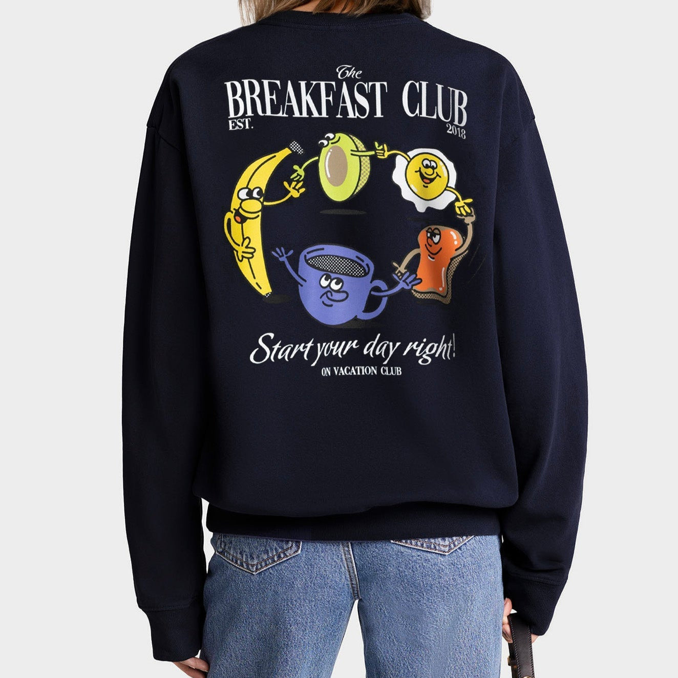 Eine Person trägt ein marineblaues Sweatshirt mit Cartoon-Frühstücksspeisen und dem Text The Breakfast Club und Start your day right! auf dem Rücken. Zu den Lebensmitteln gehören eine Banane, ein Ei, Toast und eine Kaffeetasse, die alle lächeln.
