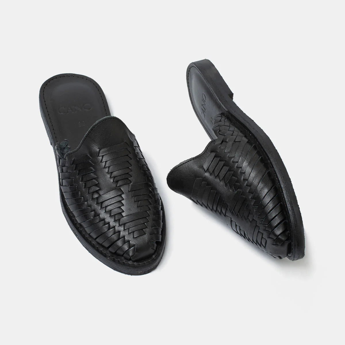 Ein Paar schwarzer geflochtener Ledersandalen, eine flach liegend, die andere auf der Seite liegend, auf einem schlichten hellgrauen Hintergrund.