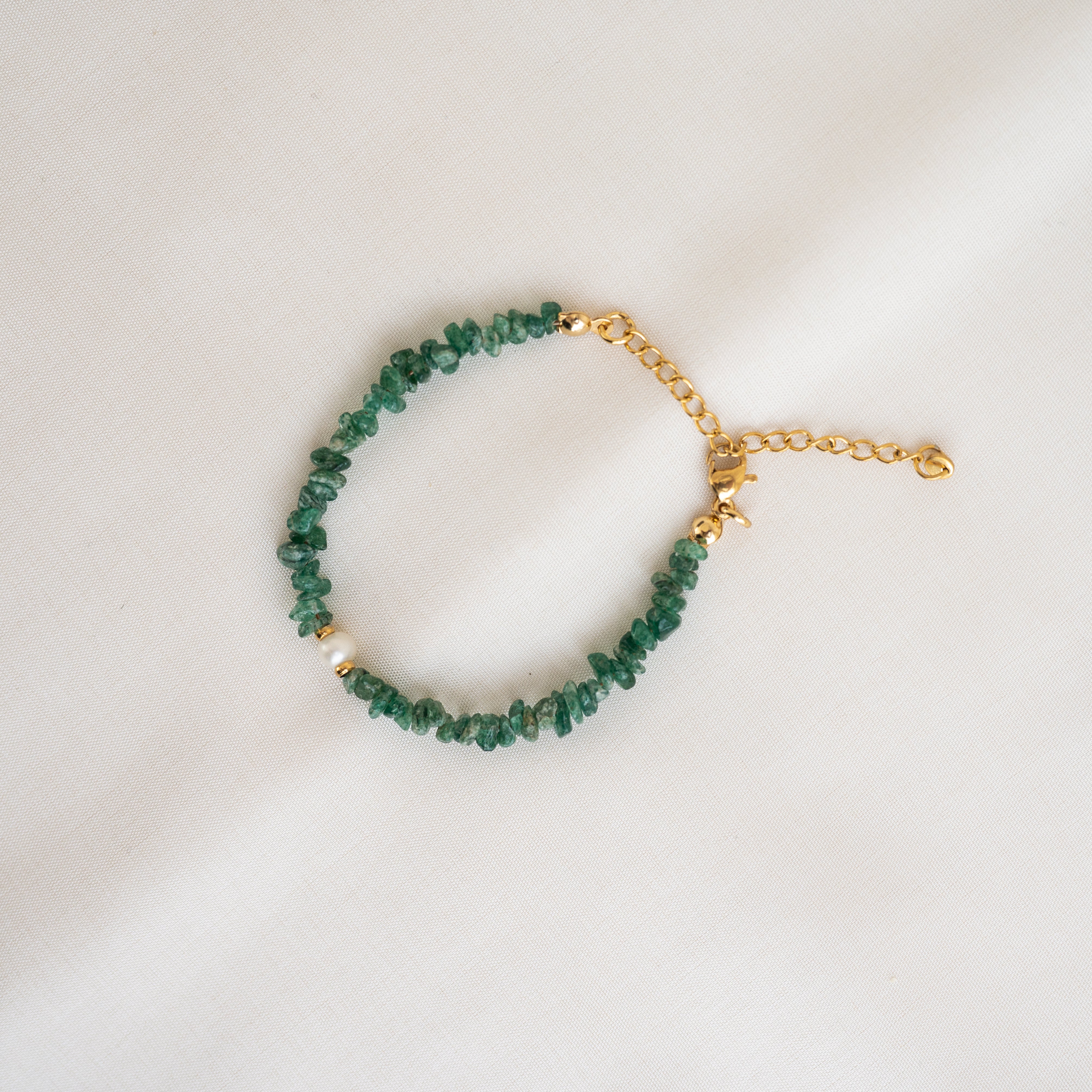 Ein goldenes Kettenarmband mit unregelmäßigen grünen Steinperlen und einem Karabinerverschluss auf einem hellbeigen Stoffhintergrund.