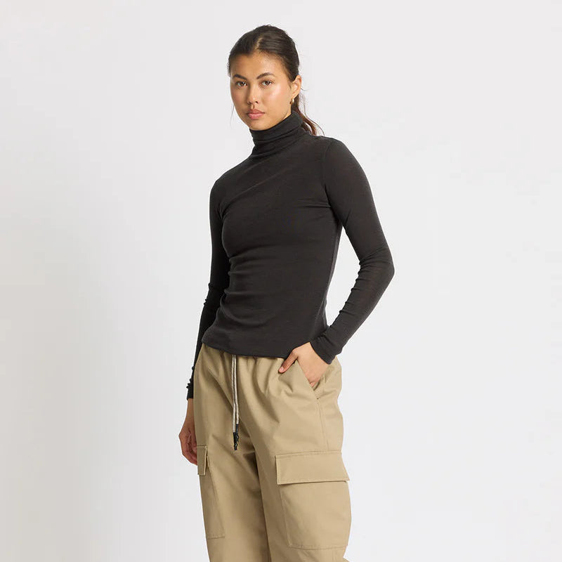 Eine Frau steht vor einem schlichten weißen Hintergrund und trägt einen schwarzen Rollkragenpullover, eine weite beigefarbene Cargohose und schwarze Adidas-Turnschuhe. Ihr Haar ist hochgesteckt, und ihre Hände stecken in den Hosentaschen.