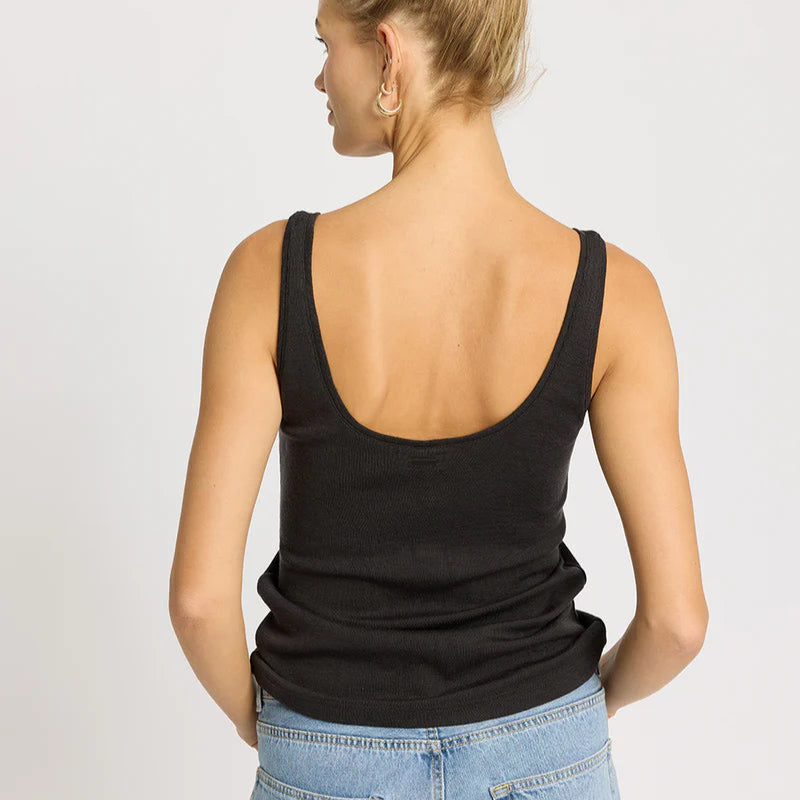 Man sieht eine Frau mit blondem Haar in einem Dutt von hinten, die ein ärmelloses schwarzes Tank-Top und hellblaue Jeans trägt. Sie steht vor einem schlichten hellen Hintergrund.