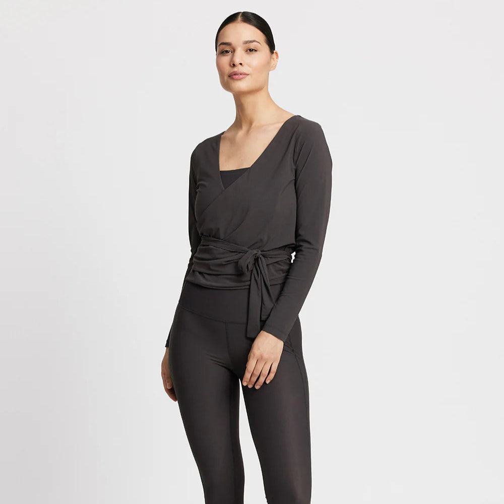 Frau in einem grauen Wickeloberteil und Leggings, vor weißem Hintergrund.