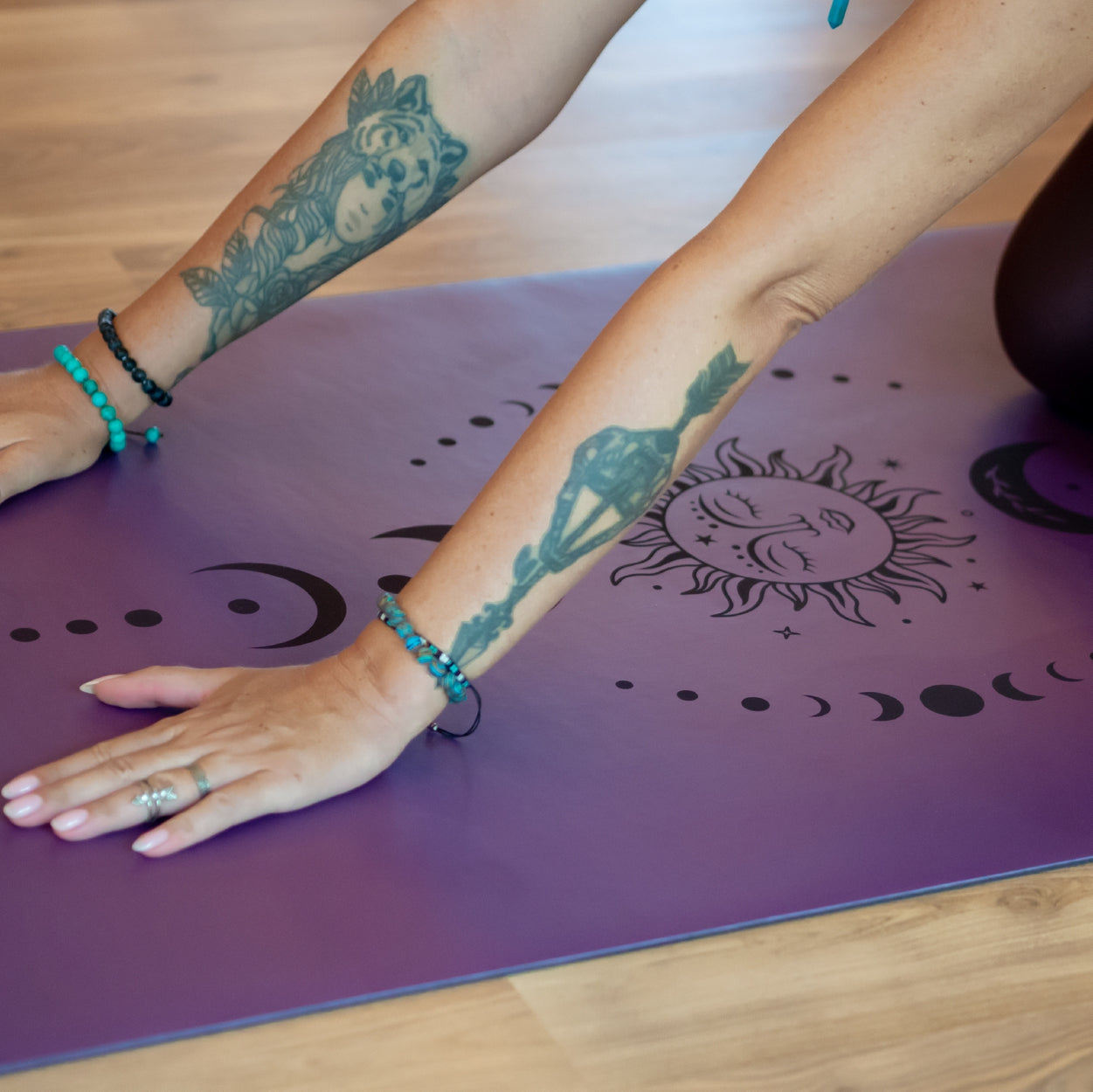 Arme mit Tattoos stützen sich auf eine lila Yogamatte mit Sonnen- und Mondmotiv, umgeben von Holzfußboden.