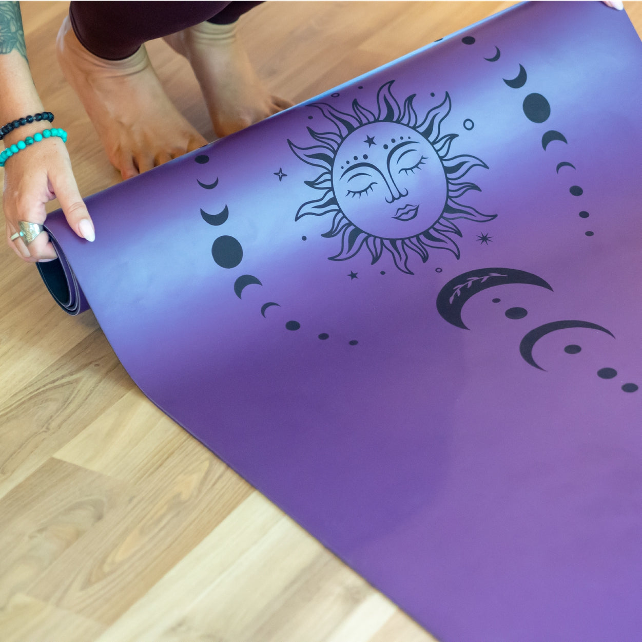 Person rolls a purple yoga mat featuring a sun and moon phase design on a wooden floor.