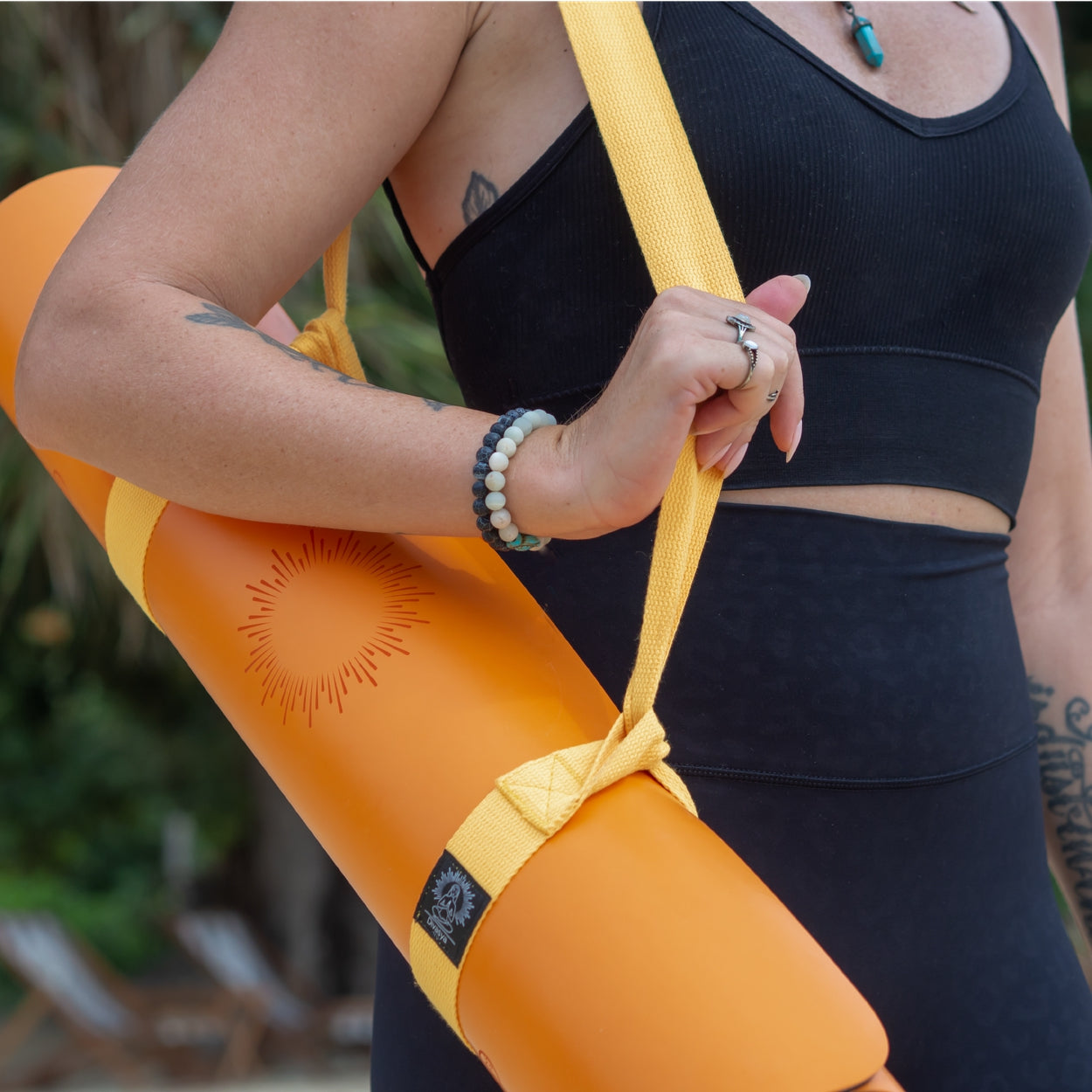 Person trägt eine orange Yogamatte mit gelbem Tragegurt und ist in Freizeitkleidung mit Armbändern.