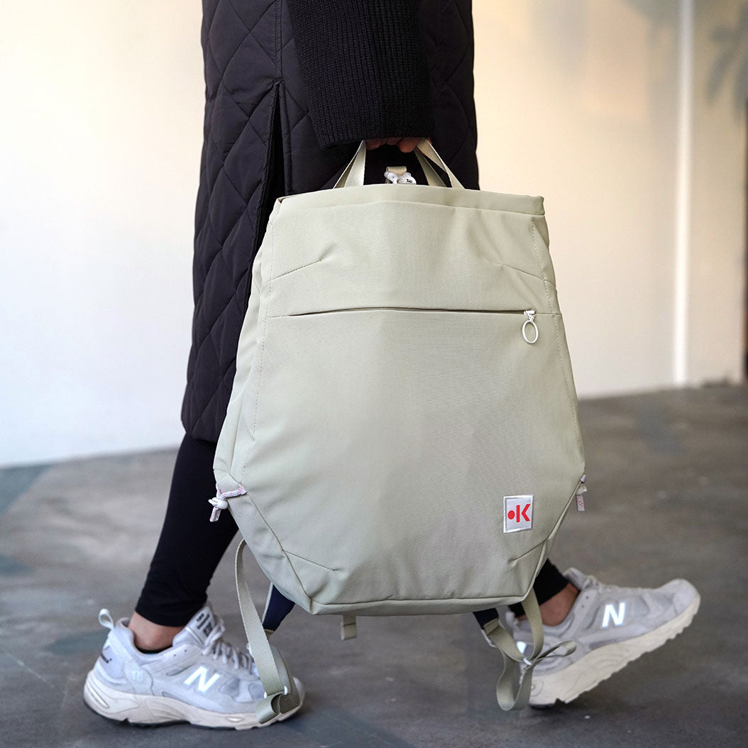 Eine Person trägt einen beigefarbenen Rucksack in der Hand, während sie Turnschuhe und einen schwarzen Mantel trägt.