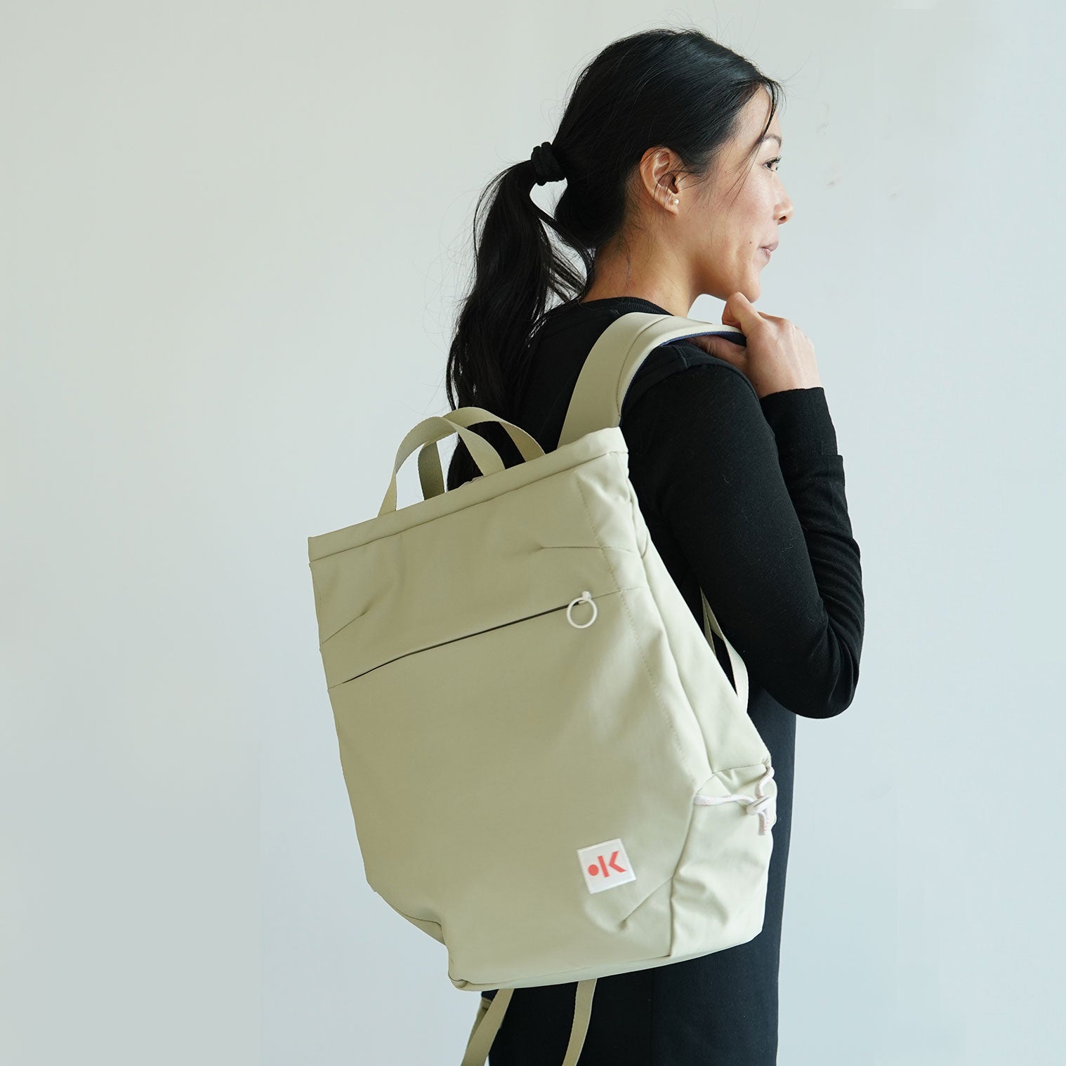 Frau mit einem beigefarbenen Rucksack und dunklem Pferdeschwanz, seitlich von hinten, vor neutralem Hintergrund.