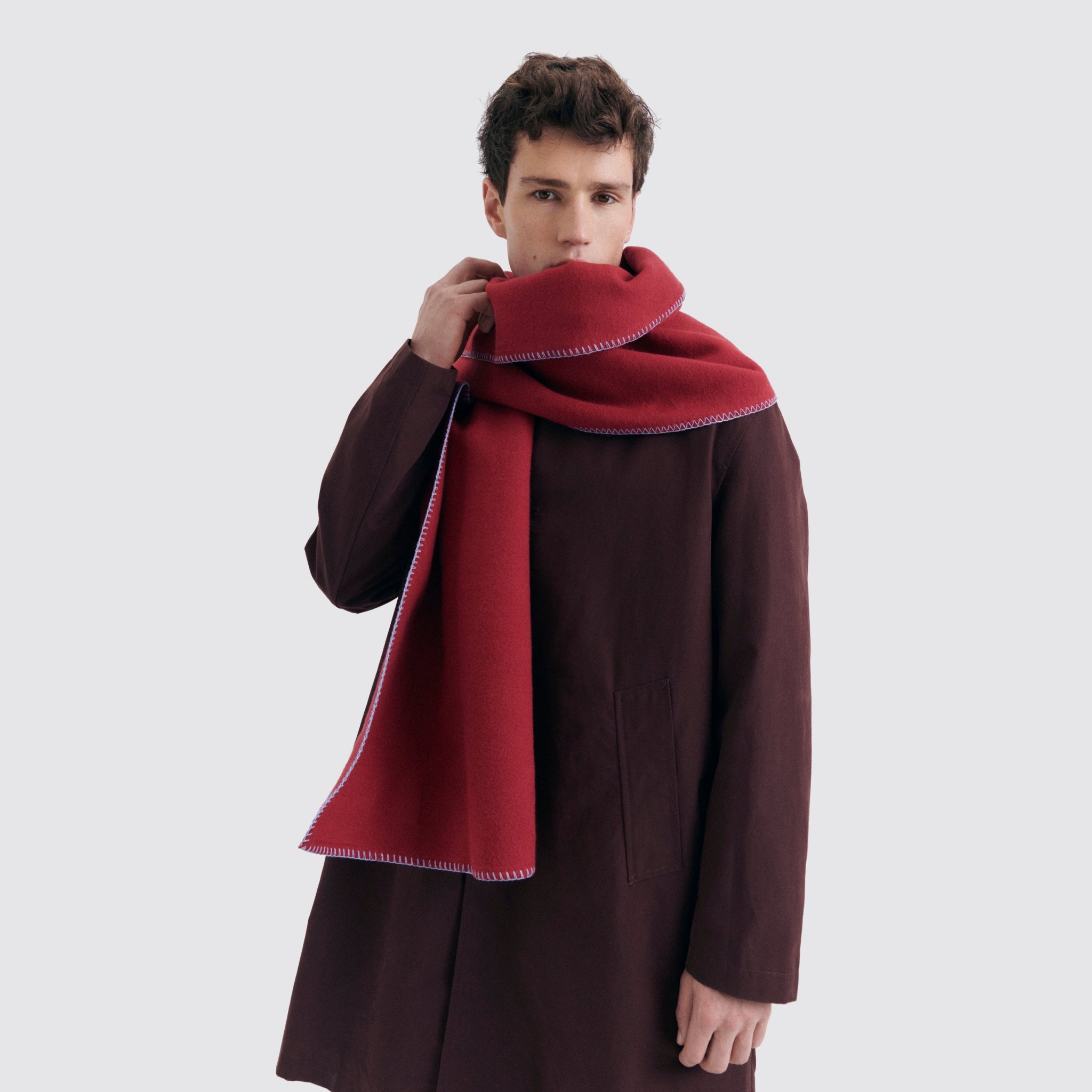 Mann in dunkelbraunem Mantel trägt einen roten Schal, der sein Gesicht teilweise bedeckt, vor grauem Hintergrund.