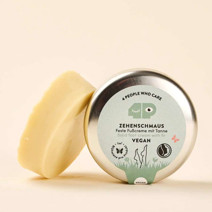 Dose mit fester Fußcreme "Zehenschmaus" neben einem Stück der Creme, beschriftet mit veganen und umweltfreundlichen Symbolen.