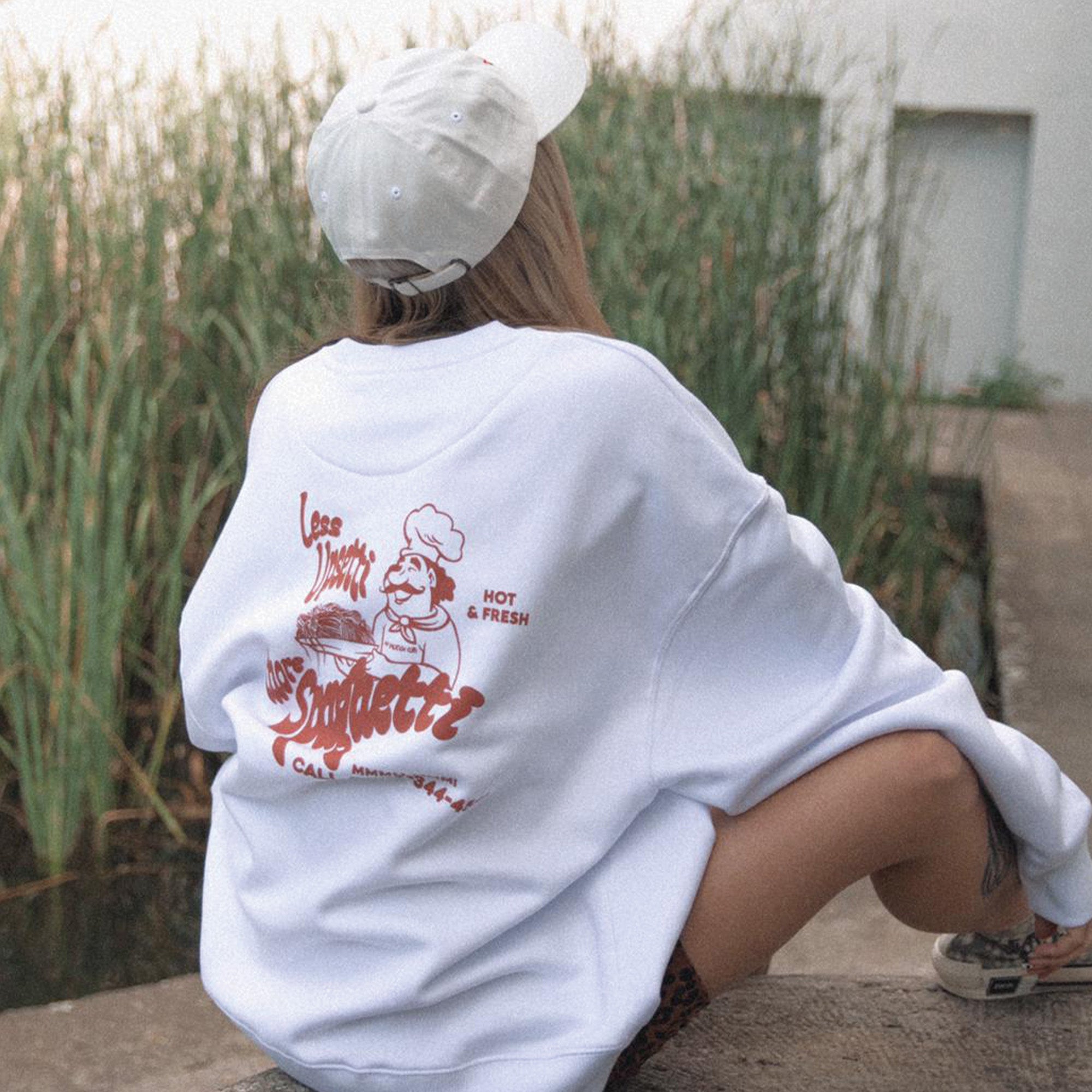 Eine Person, die eine weiße Mütze und ein übergroßes weißes Sweatshirt mit einer roten Grafik auf dem Rücken trägt, sitzt auf einem Betonweg in der Nähe von hohem Gras und blickt von der Kamera weg.
