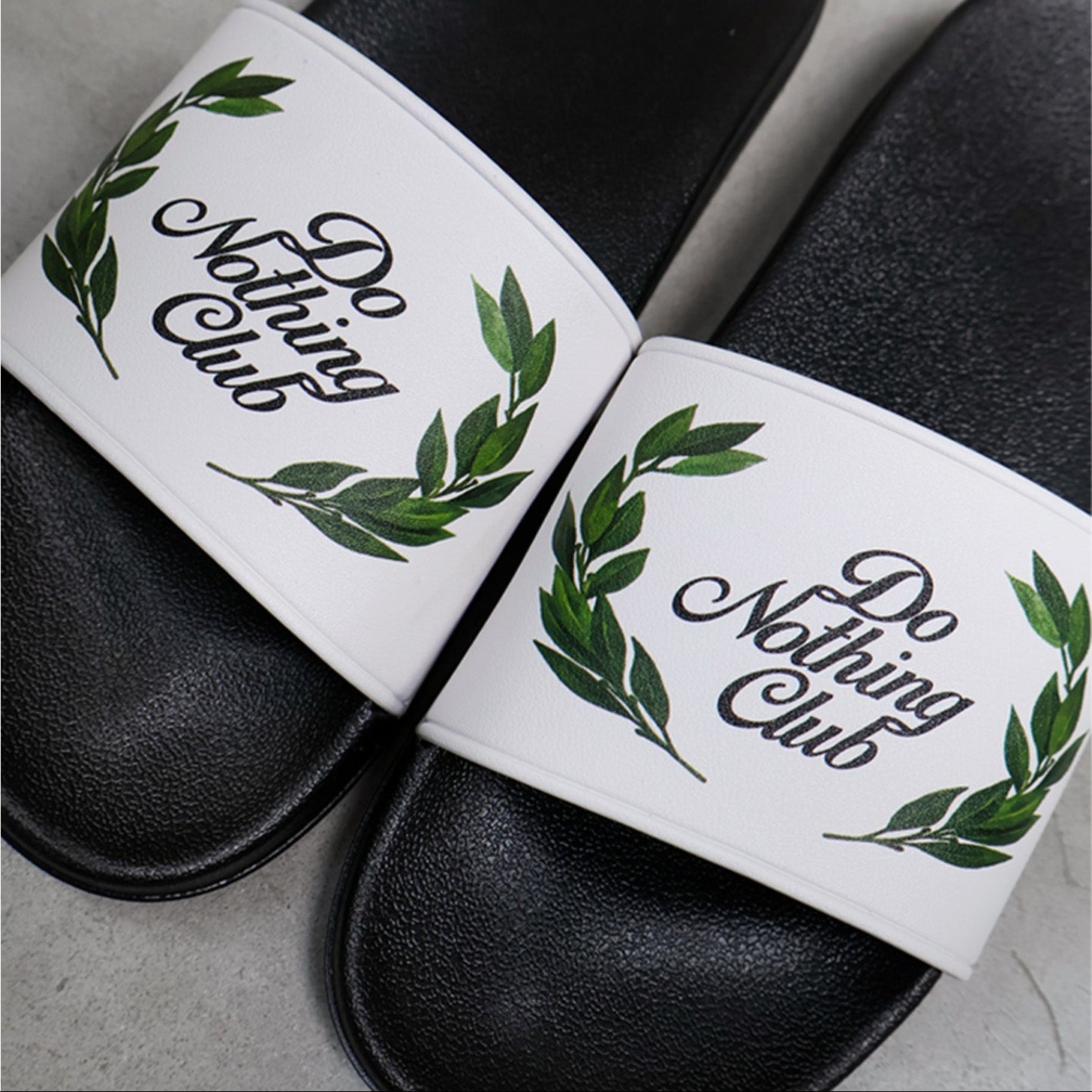 Eine Nahaufnahme von schwarzen Slide-Sandalen mit weißen Riemen, auf denen in Kursivschrift "Do Nothing Club" steht, umgeben von grünen Blattkränzen. Die Sandalen sind nebeneinander auf einer hellgrauen Fläche platziert.
