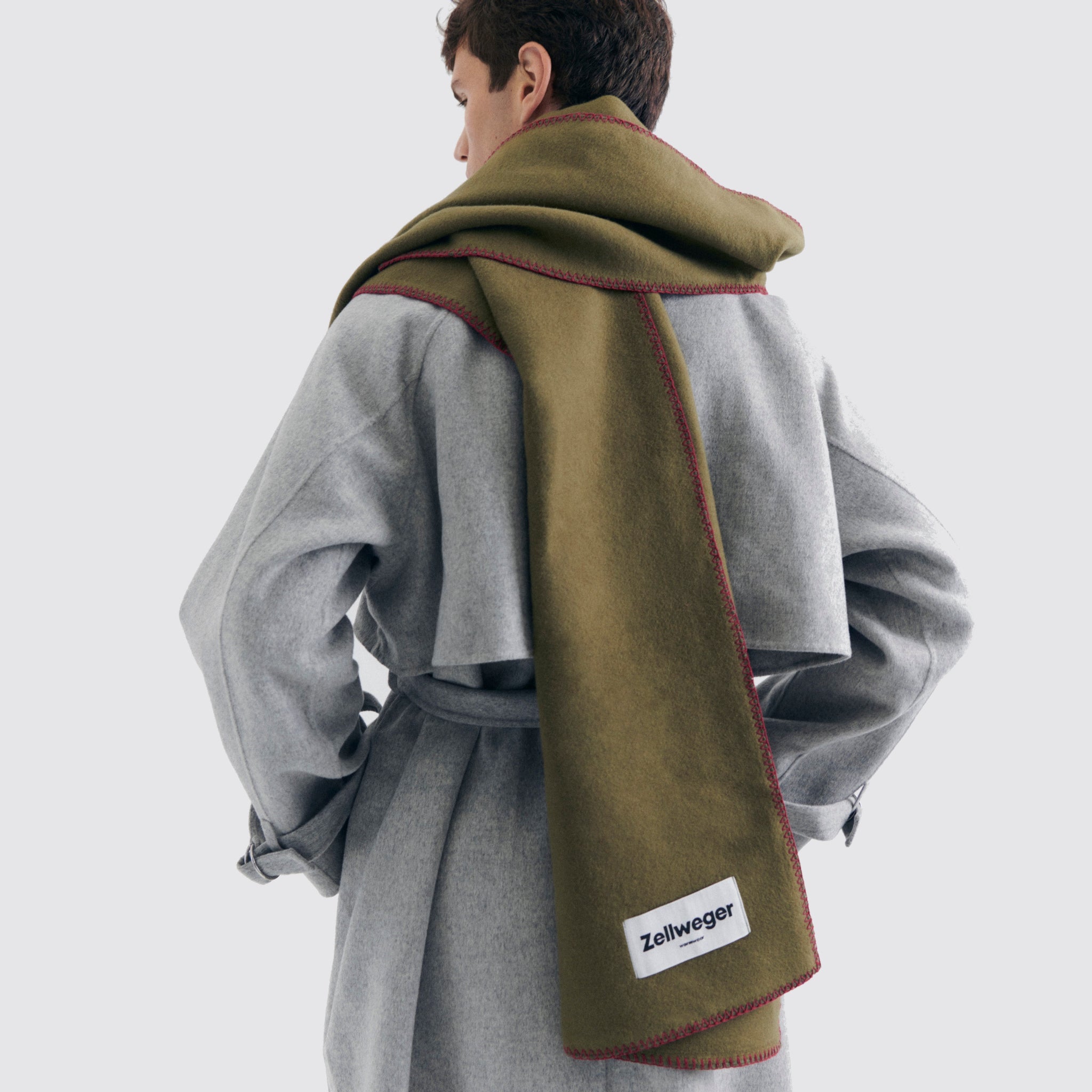 Person in einem grauen Mantel mit einem langen, olivgrünen Schal mit Label "Zellweger" auf hellem Hintergrund.