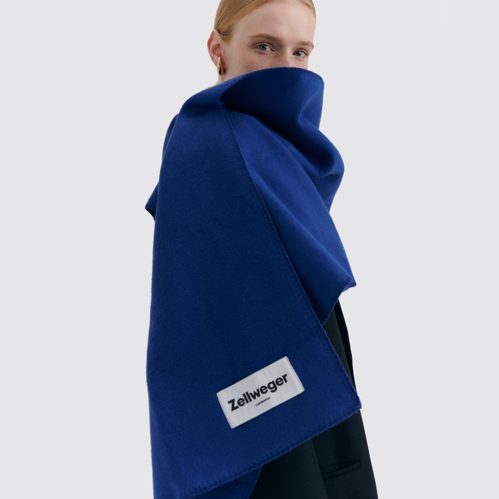 Eine Person trägt einen blauen Schal mit dem Etikett "Zellweger" vor hellem Hintergrund.