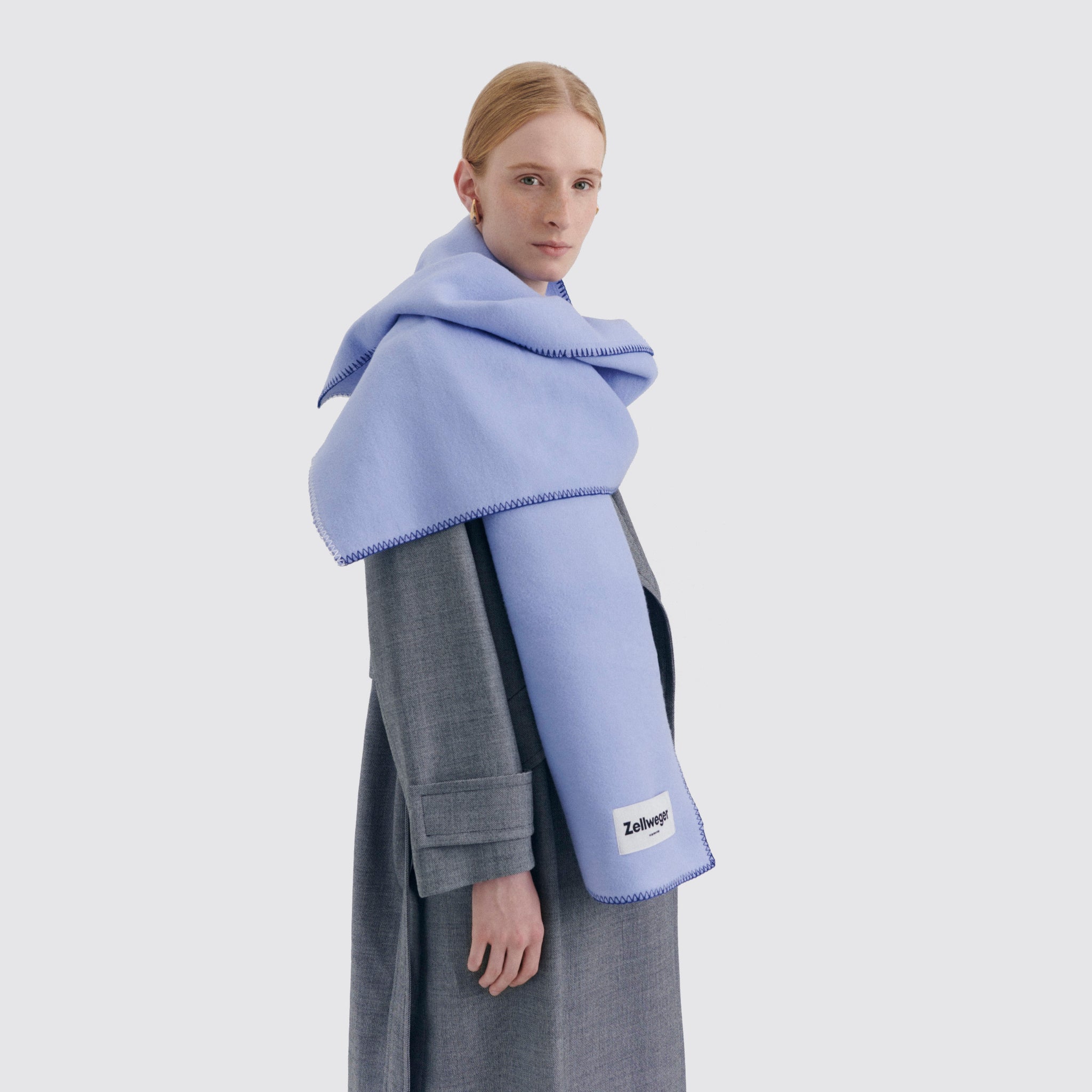 Person in grauem Mantel trägt einen großen, blauen Schal mit einem weißen Etikett, vor neutralem Hintergrund.