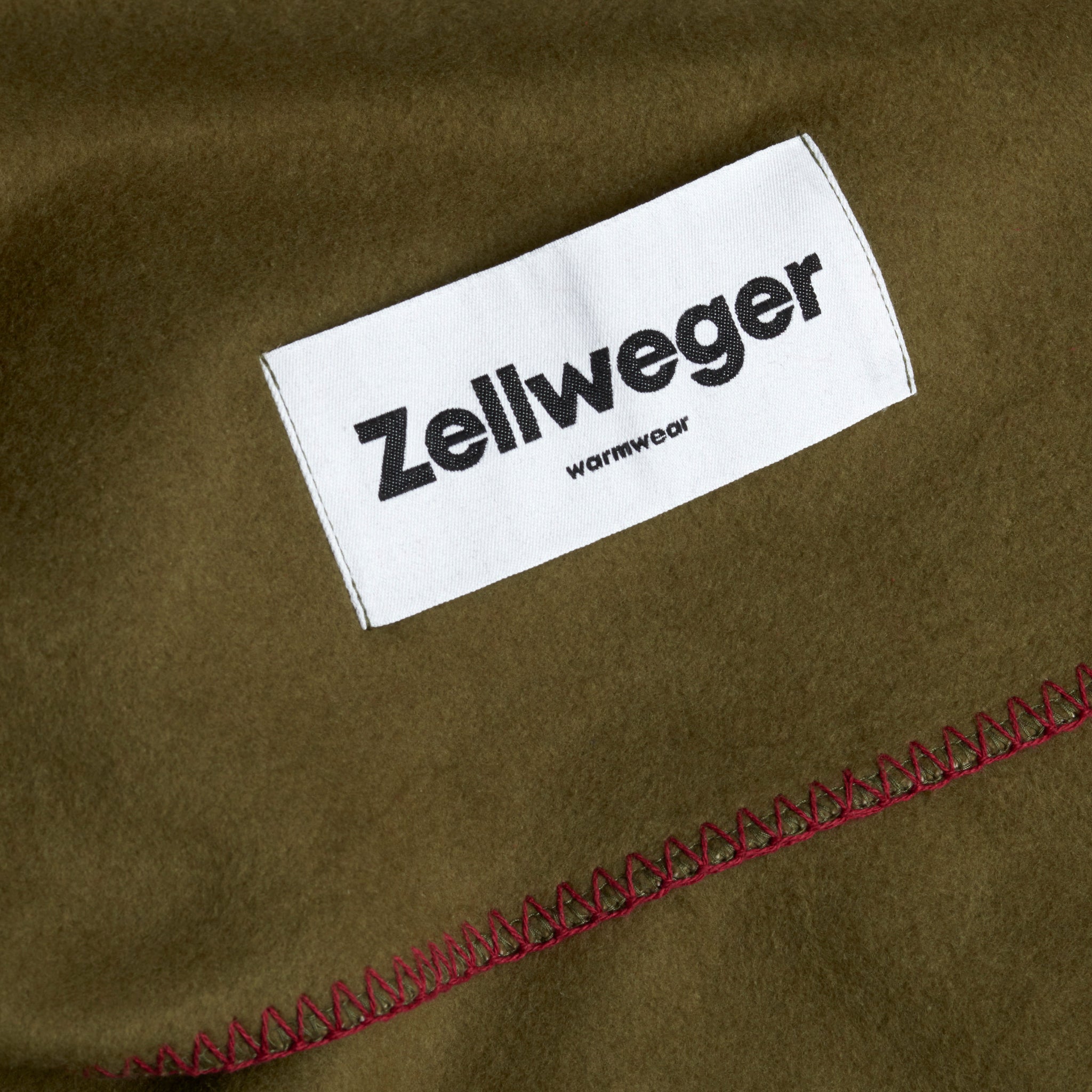 Olivefarbener Stoff mit roter Naht und weißem Etikett, das "Zellweger warmwear" zeigt.