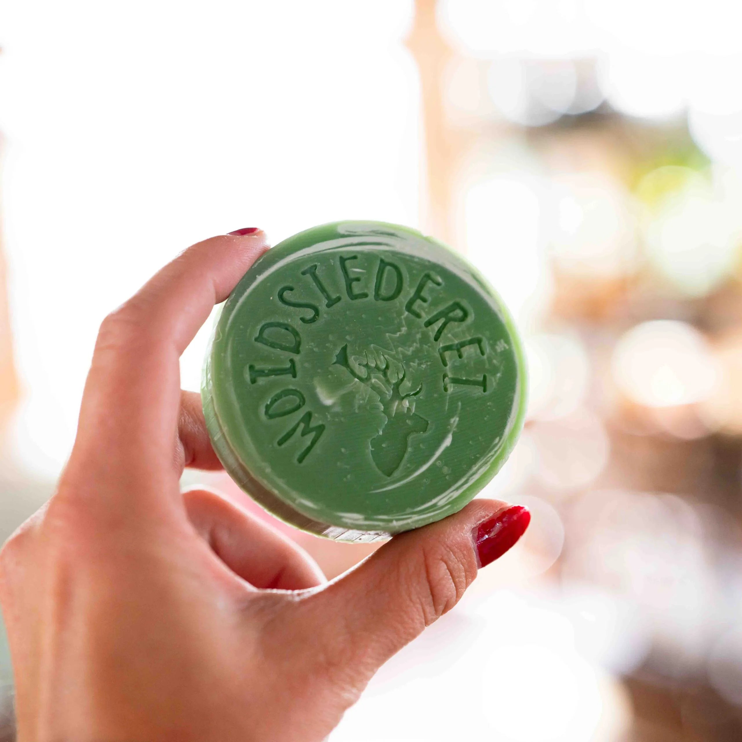 Eine Hand mit rot lackierten Nägeln hält ein rundes grünes Seifenstück mit der Aufschrift WOLDSIEDEREI und einem Hirschkopf-Logo in der Hand. Der Hintergrund ist sanft verschwommen.