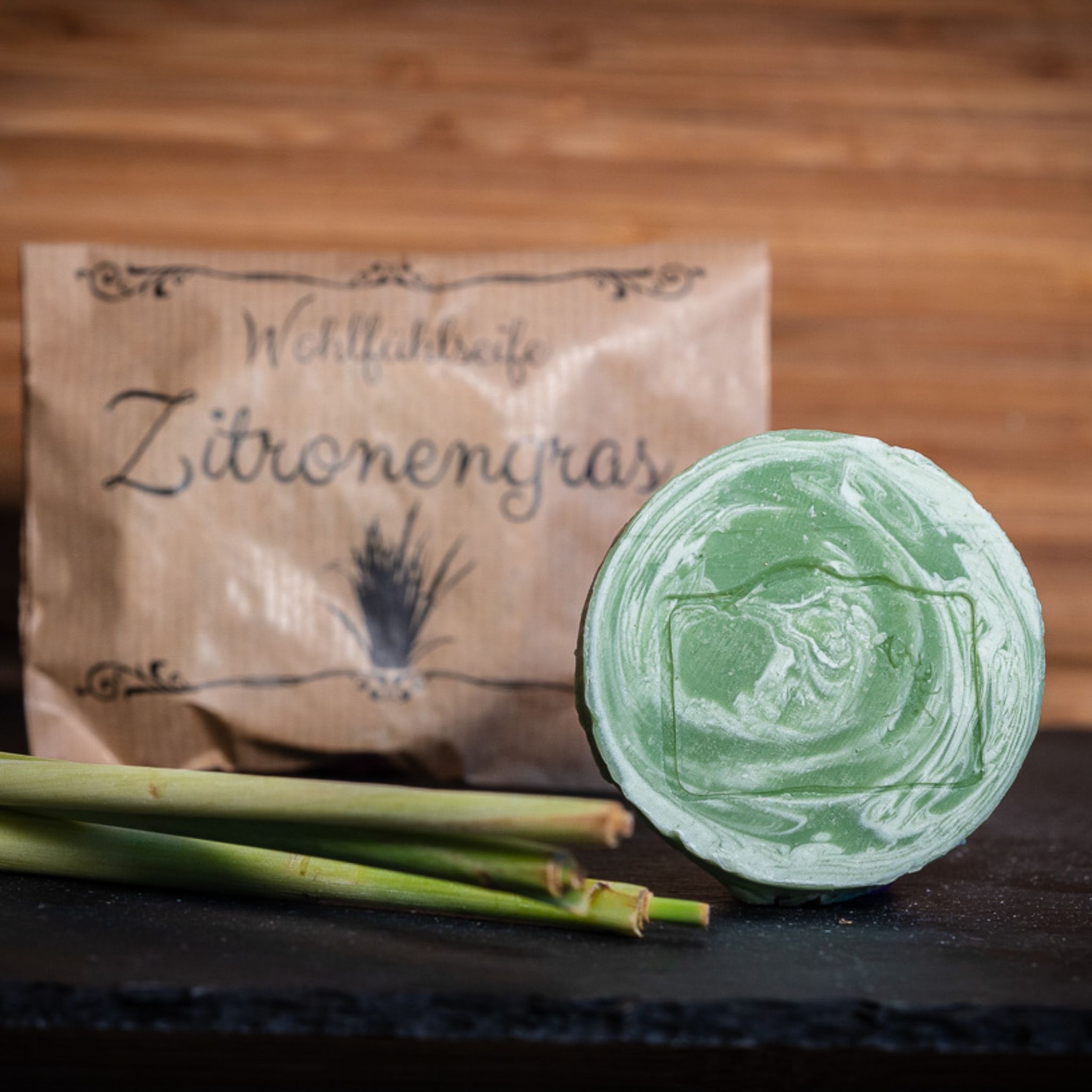 Runde grüne Seife vor einer Verpackung mit der Aufschrift "Wohlfühlseife Zitronengras", daneben Zitronengrasstangen.