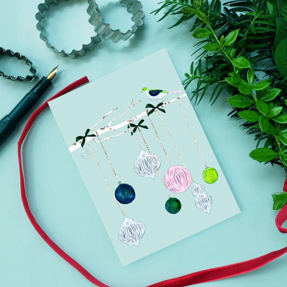 Eine festliche Grußkarte mit bunten Ornamenten, die von einem weißen Zweig herabhängen, auf einer hellblauen Fläche neben einem roten Band, grünem Laub, einem Stift und Ausstechformen aus Metall.