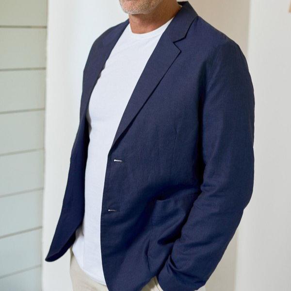 Person in einem dunkelblauen Sakko mit weißem T-Shirt, steht vor einer hellen Wand.