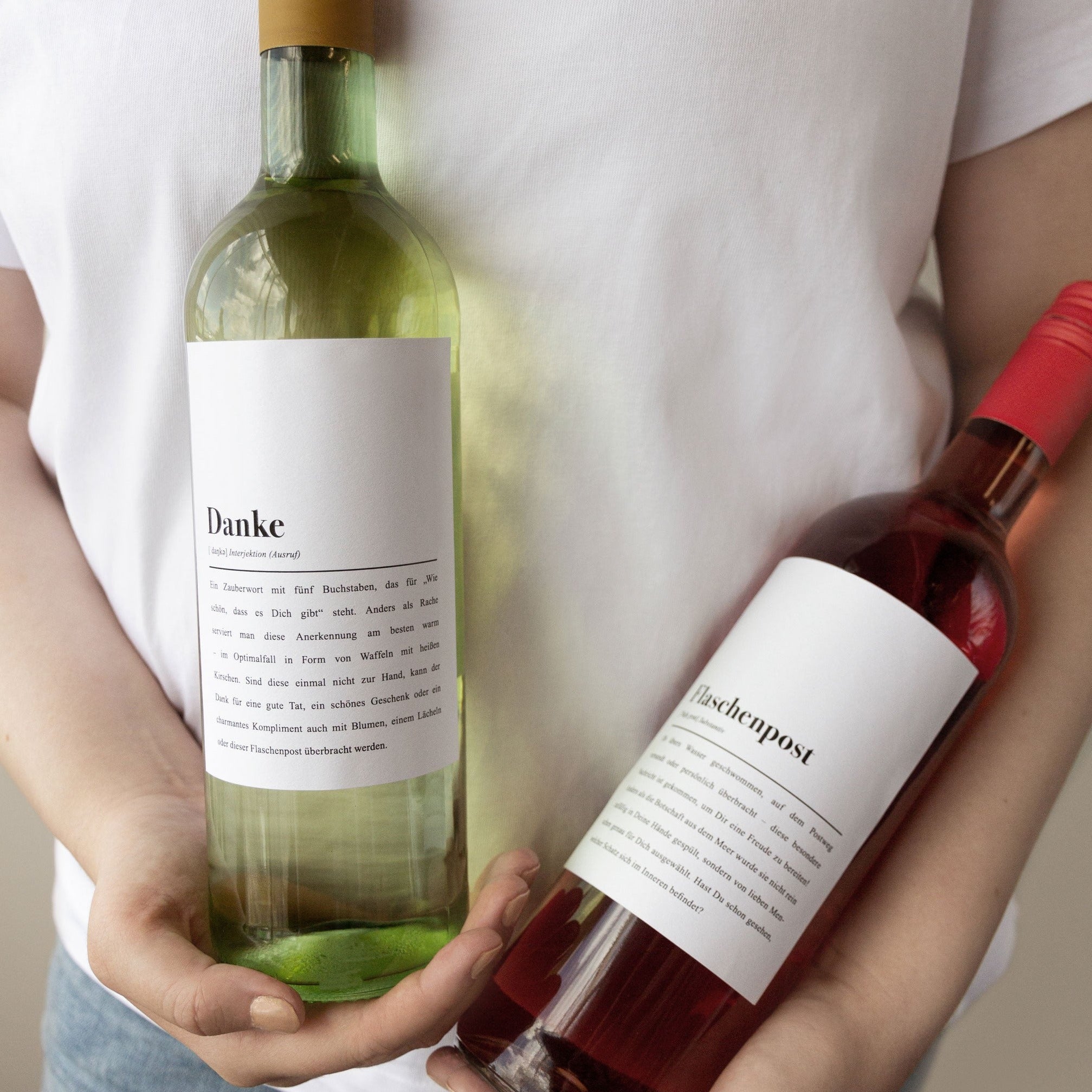 Person hält zwei Weinflaschen mit weißen Etiketten, eine grüne und eine rote, beschriftet mit "Danke" und "Flaschenpost".