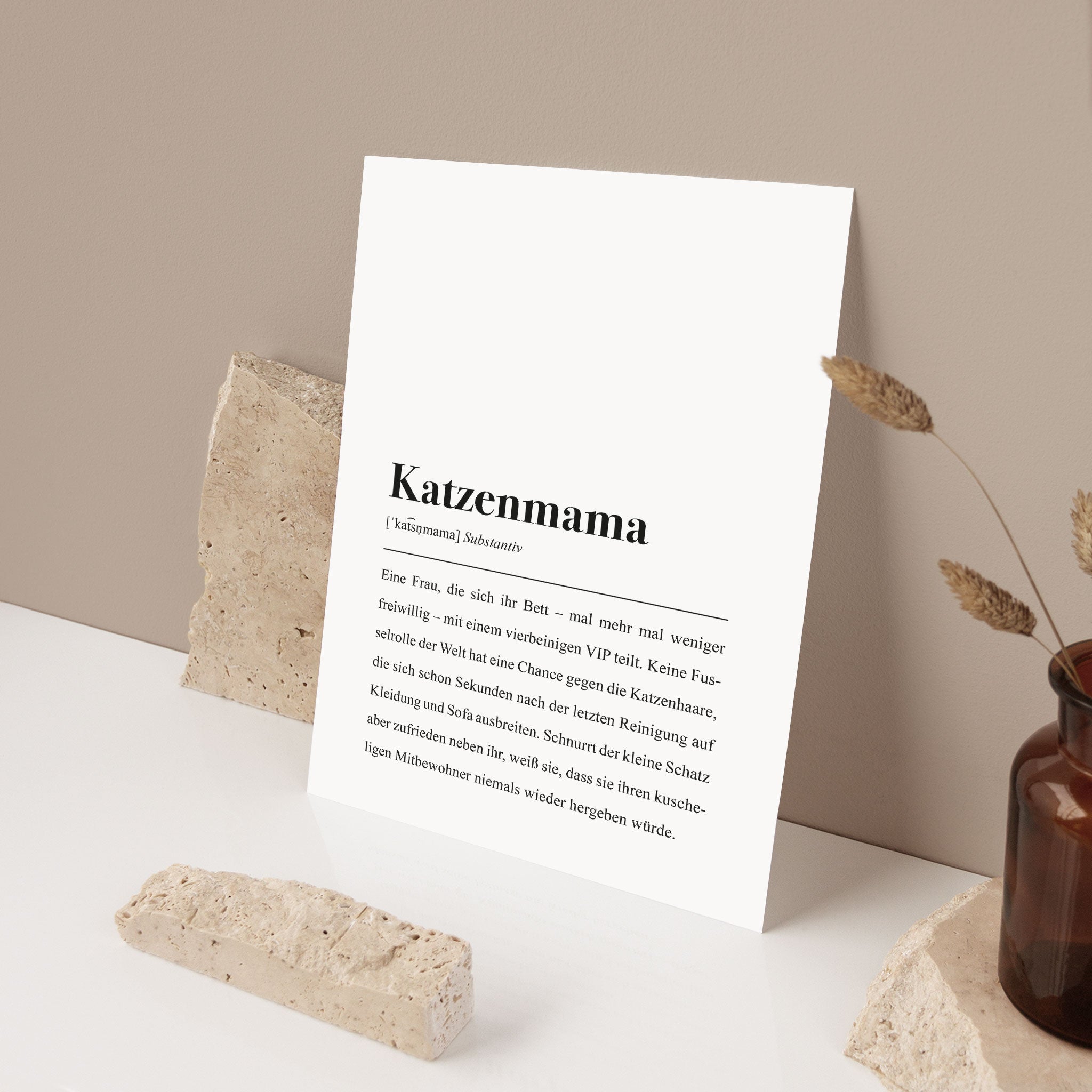 Ein Schild mit der Definition von "Katzenmama", aufgerichtet zwischen zwei Steinen, daneben eine Vase mit Zweigen.