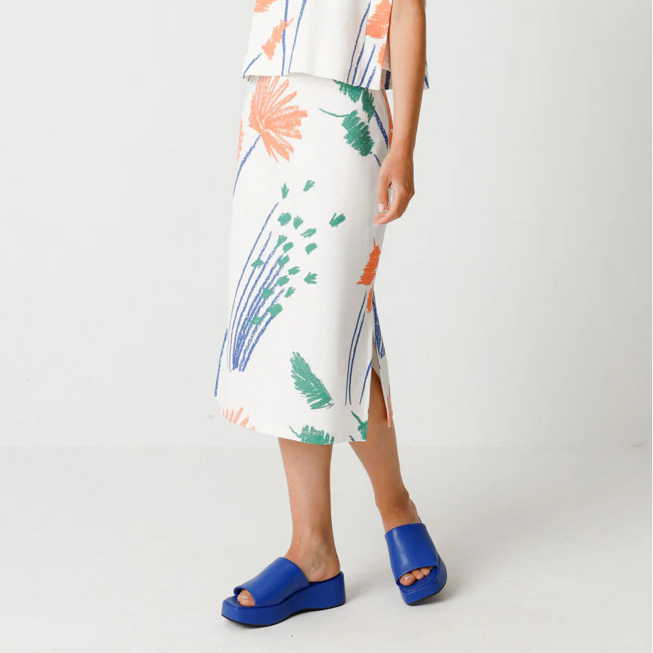 Eine Frau steht vor einem weißen Hintergrund und trägt ein ärmelloses, lockeres weißes Oberteil und einen passenden Midirock mit einem orangefarbenen, blauen und grünen Blumenmuster. Sie blickt nach unten und trägt hellblaue Plateausandalen.