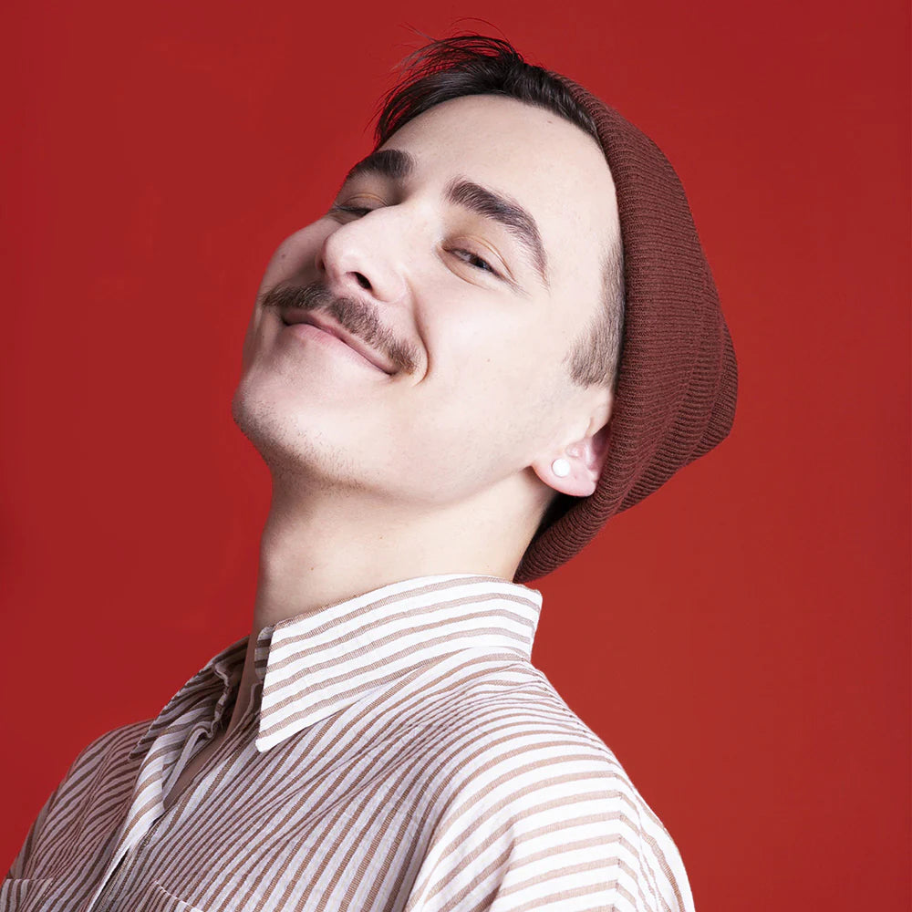 Eine lächelnde Person mit Schnurrbart, die eine braune Mütze und ein beigefarbenes gestreiftes Hemd trägt, posiert vor einem einfarbig roten Hintergrund.