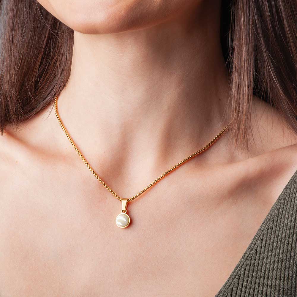 Eine Frau trägt eine goldene Halskette mit einem runden Anhänger. Sie hat glattes braunes Haar und ist mit einem olivgrünen, schulterfreien Oberteil bekleidet. Der Hintergrund ist neutral.