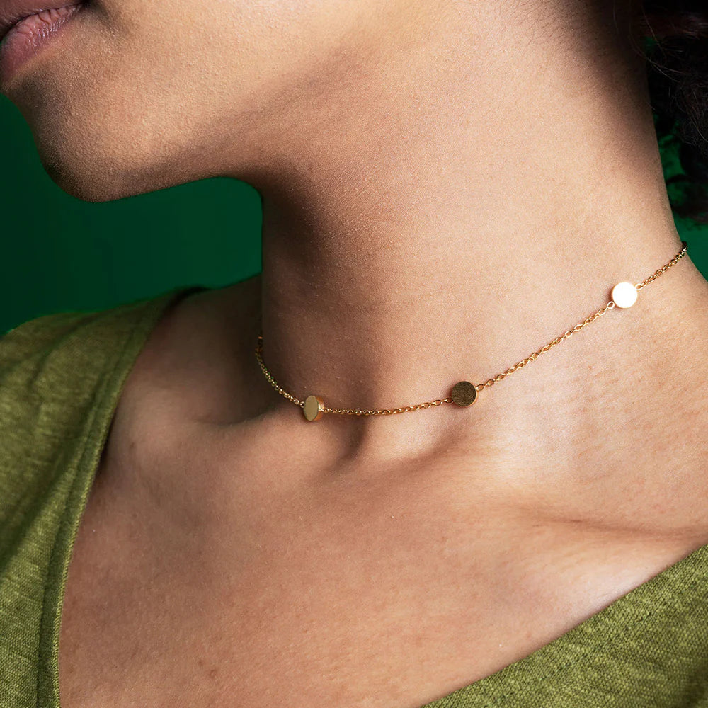 Nahaufnahme einer Person, die eine goldene Kettenhalskette mit kleinen runden Scheiben vor einem grünen Hintergrund trägt. Die Person ist mit einem grünen Oberteil bekleidet.