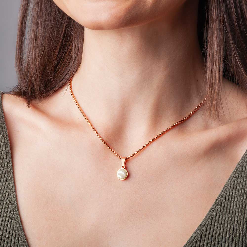 Eine Frau trägt eine goldene Halskette mit einem runden, goldenen Anhänger. Sie hat glattes braunes Haar und ist mit einem olivgrünen Oberteil mit tiefem V-Ausschnitt bekleidet. Der Schwerpunkt des Bildes liegt auf ihrem Hals und ihrem Schlüsselbeinbereich.