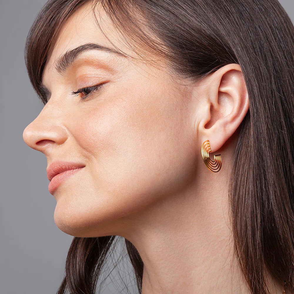 Eine Frau mit glattem braunem Haar ist im Profil dargestellt und trägt einen goldenen, modernen, gestuften Reif-Ohrring. Sie hat ein sanftes Make-up und ein leichtes Lächeln, vor einem schlichten grauen Hintergrund.