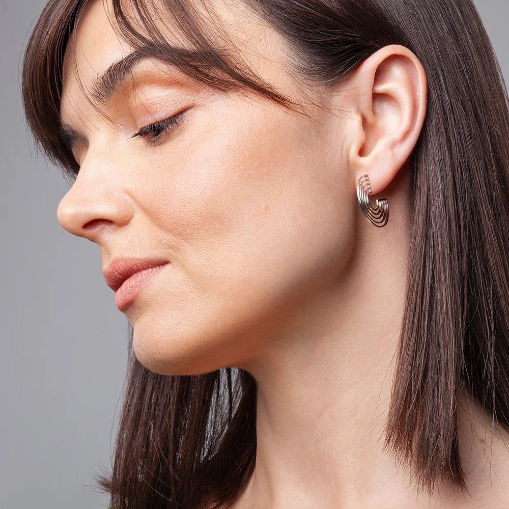 Eine Frau mit glattem braunem Haar und natürlichem Make-up ist im Profil dargestellt und trägt einen klobigen, gedrehten Reif-Ohrring. Der Hintergrund ist einfarbig grau.