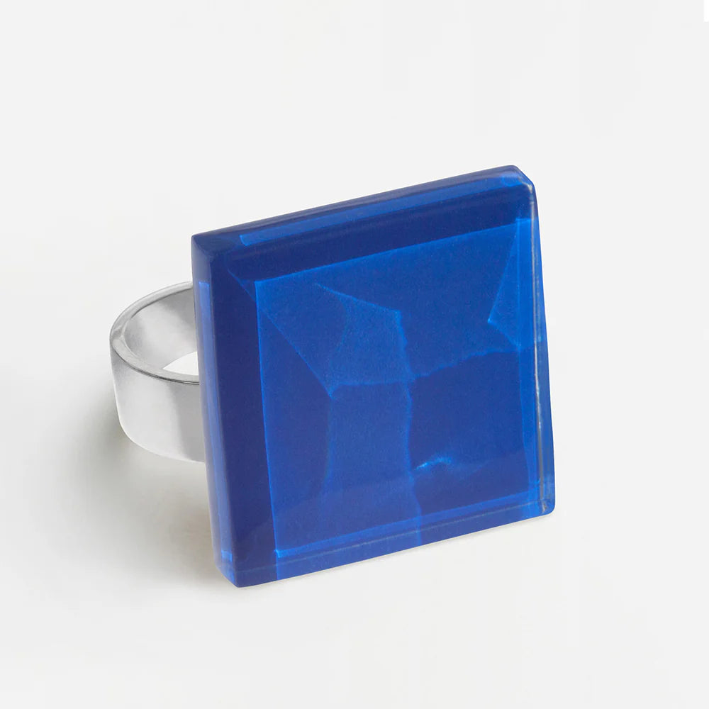 Ein Silberring mit einem großen, quadratischen, facettierten blauen Edelstein mit modernem, geometrischem Design vor einem schlichten weißen Hintergrund.