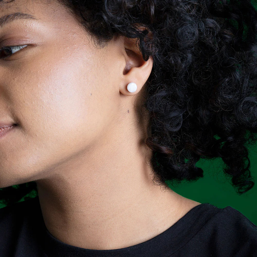 Nahaufnahme einer Person mit dunklem lockigem Haar, die einen runden weißen Ohrstecker im Ohr trägt, vor einem grünen Hintergrund. Nur ein Teil des Gesichts und des Ohrs ist sichtbar.