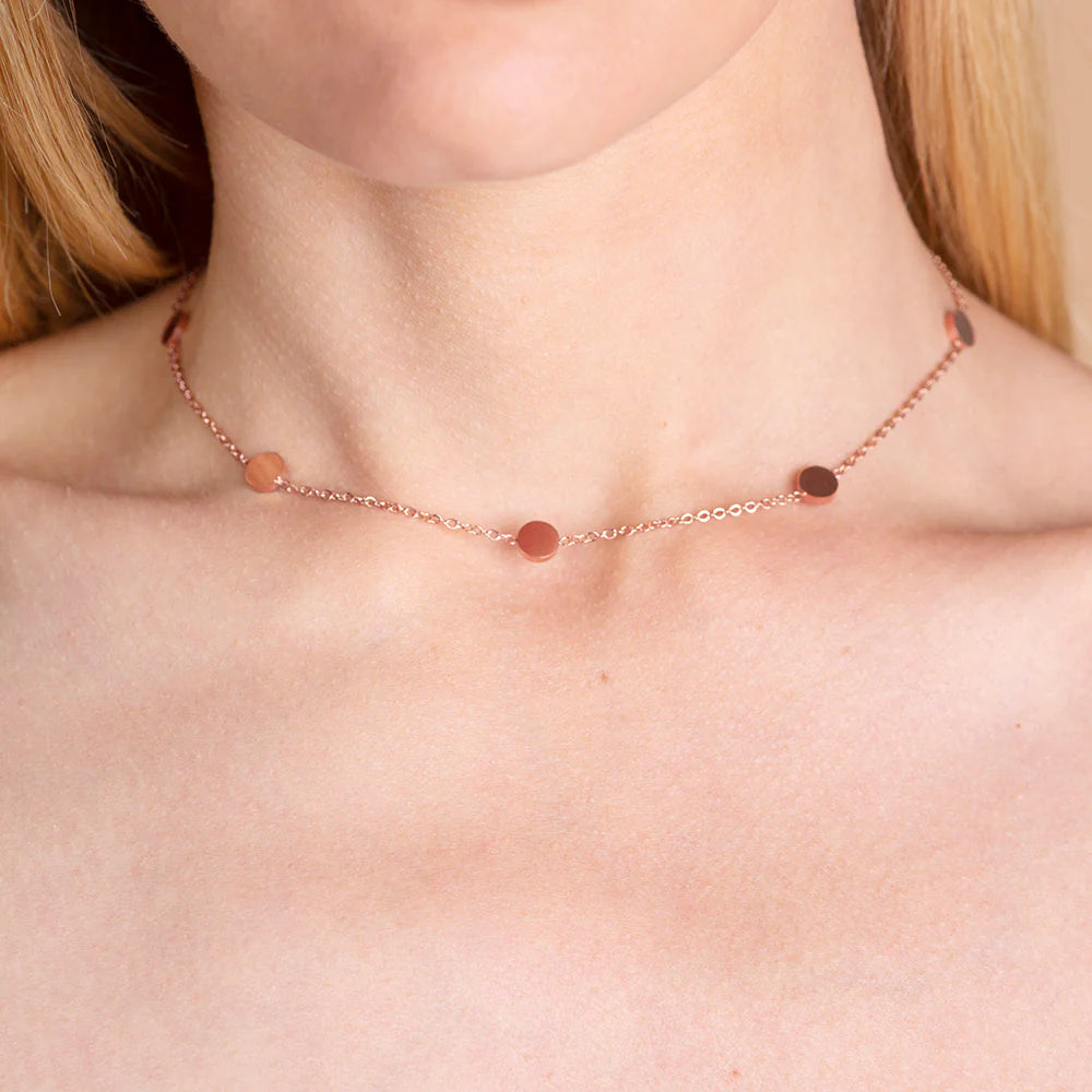 Nahaufnahme einer Person, die eine zarte roségoldene Halskette mit kleinen, gleichmäßig verteilten runden rosa und braunen Perlen trägt. Die Halskette sitzt knapp oberhalb des Schlüsselbeins.