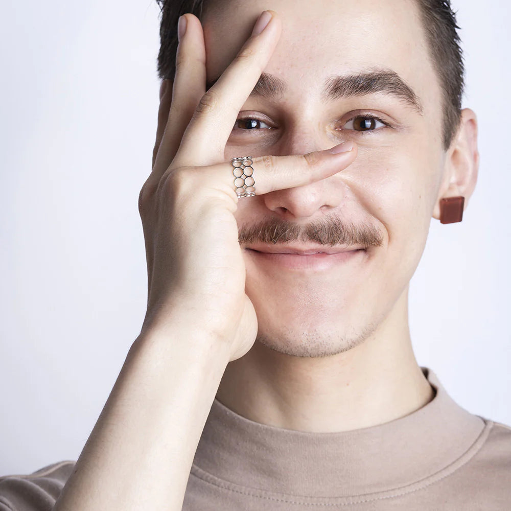 Eine lächelnde Person mit Schnurrbart hält ihre Hand über einen Teil ihres Gesichts und formt mit ihren Fingern einen Kreis um ein Auge. Sie trägt einen geometrischen Ring und einen quadratischen Ohrring und ist mit einem leichten, hochgeschlossenen Hemd bekleidet.