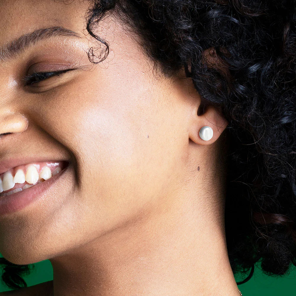 Nahaufnahme einer lächelnden Frau mit lockigem Haar, die an ihrem linken Ohr einen kleinen, runden, weißen Ohrstecker trägt. Der Hintergrund ist grün.