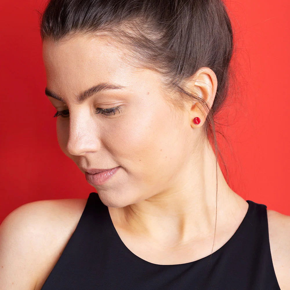 Eine junge Frau mit braunen Haaren in einem Pferdeschwanz blickt nach unten. Sie trägt ein schwarzes ärmelloses Oberteil und kleine rote Ohrstecker vor einem einfarbig roten Hintergrund.