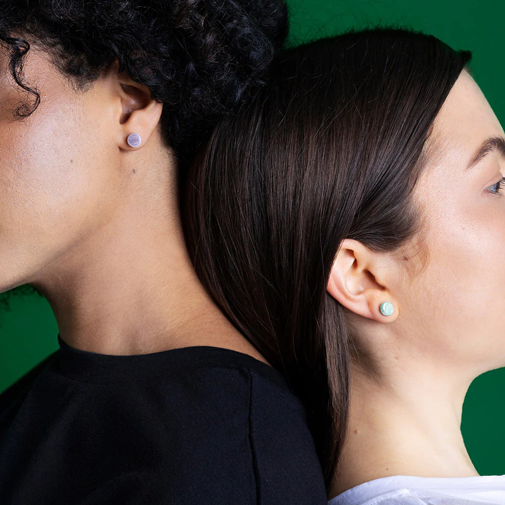 Zwei Personen stehen Rücken an Rücken vor einem grünen Hintergrund, beide tragen Ohrstecker. Eine Person hat lockiges dunkles Haar und ein schwarzes Oberteil, die andere hat glattes braunes Haar und ein weißes Hemd.