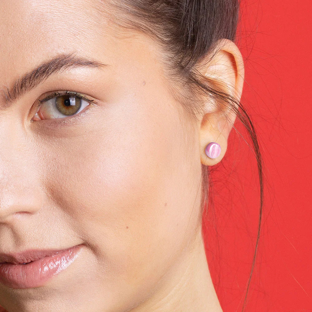 Nahaufnahme einer Frau vor einem roten Hintergrund, die die rechte Gesichtshälfte zeigt. Sie trägt einen kleinen, runden, rosafarbenen Ohrstecker und hat eine glatte, klare Haut.