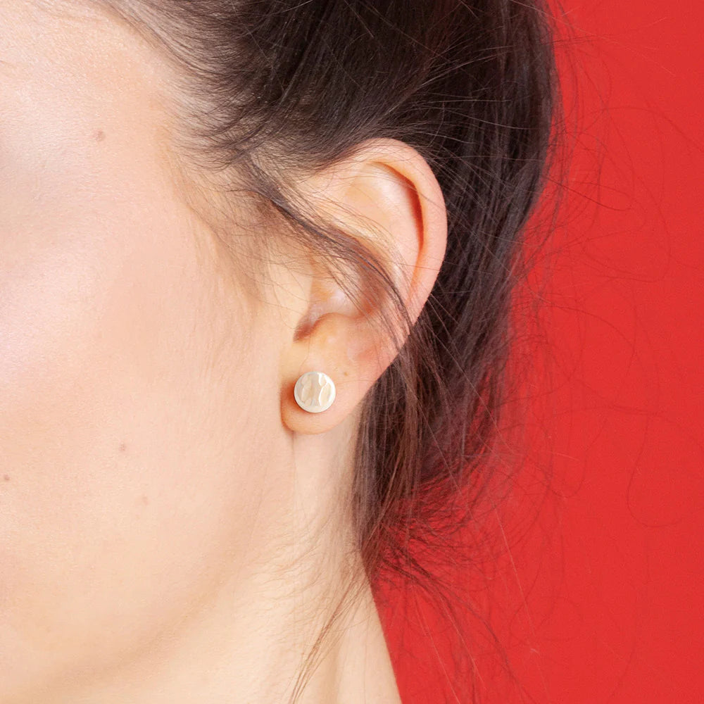 Nahaufnahme eines Frauenohrs, das einen kleinen, runden, hellen Ohrstecker trägt. Der Hintergrund ist einfarbig rot, und ein Teil ihres braunen Haars ist sichtbar.