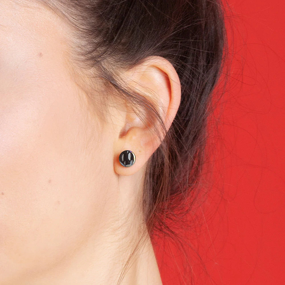 Nahaufnahme des Ohrs einer Person, die einen kleinen, runden schwarzen Ohrstecker trägt. Die Person hat helle Haut und dunkles, zurückgebundenes Haar mit rotem Hintergrund.