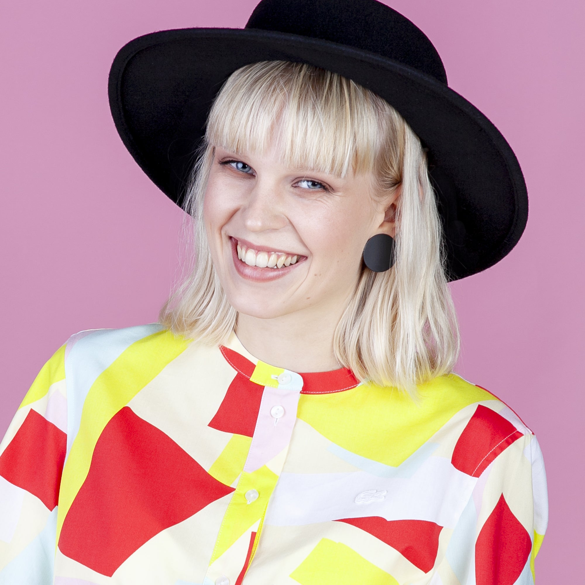 Eine lächelnde Person mit blondem Haar trägt einen breitkrempigen schwarzen Hut, runde schwarze Ohrringe und ein buntes, geometrisch gemustertes Hemd und steht vor einem rosa Hintergrund.