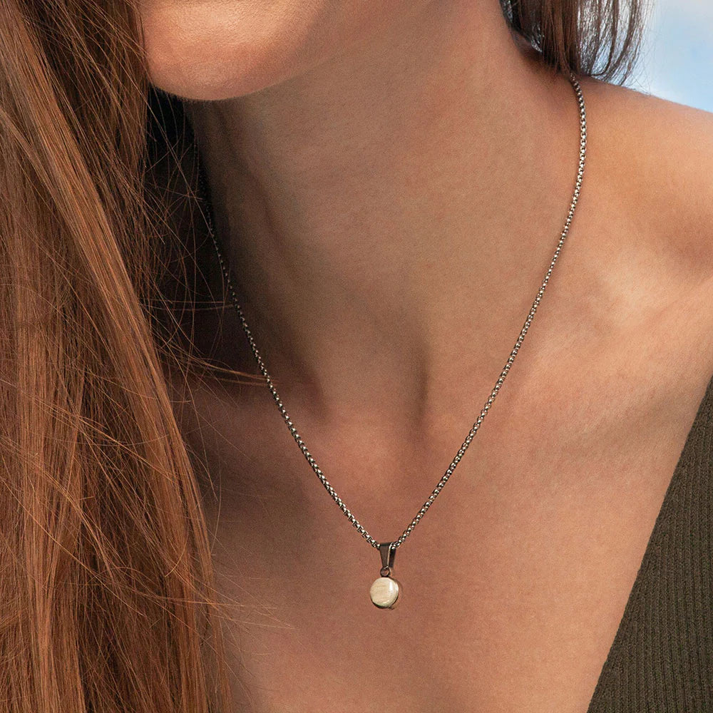 Eine Frau, die eine silberne Halskette mit einem runden weißen Anhänger trägt, wird von den Schultern aufwärts gezeigt. Sie hat langes braunes Haar und trägt ein dunkles, ärmelloses Oberteil. Der Hintergrund ist sanft verschwommen.