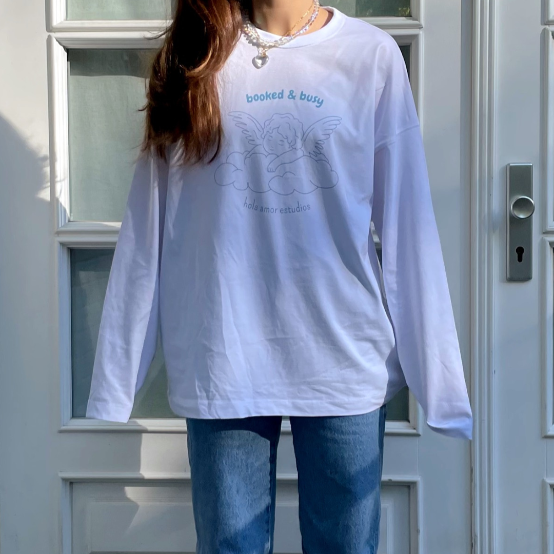 Eine Person, die ein lockeres, weißes, langärmeliges Hemd mit einem gebuchten und beschäftigten Engelsmuster, blaue Jeans und eine Kettenhalskette trägt, steht vor einer weißen Tür mit Glasscheiben.