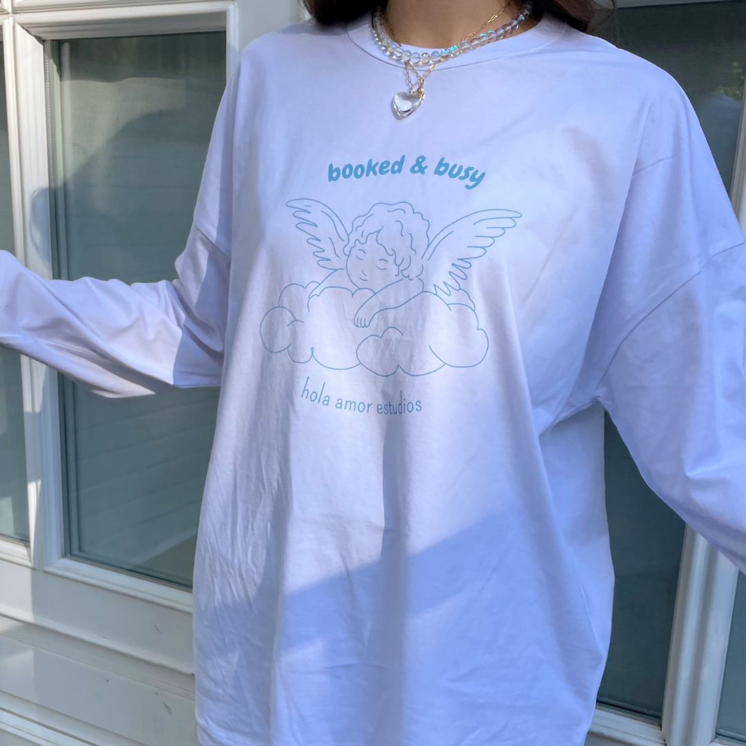 Eine Person trägt ein weißes langärmeliges Hemd mit dem Umriss eines auf Wolken ruhenden Engels und dem Text booked & busy und hola amor studios. Die Person trägt außerdem eine mehrschichtige silberne Halskette.