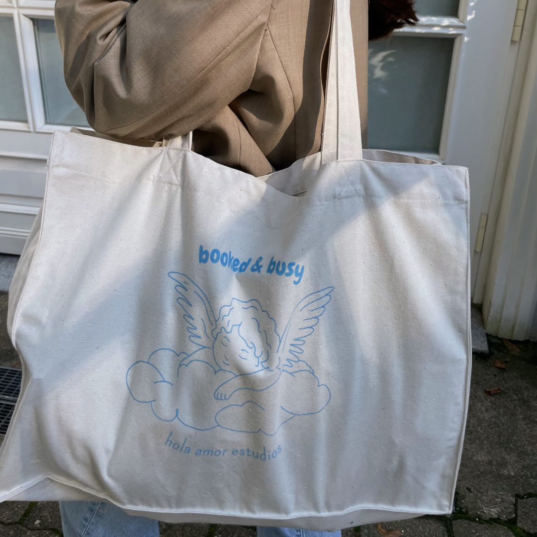 Eine Person trägt eine beigefarbene Tragetasche mit einer Illustration eines geflügelten Cherubs, der auf einer Wolke ruht. Der Text auf der Tasche lautet booked & busy und hola amor estudios. Die Person trägt eine braune Jacke.