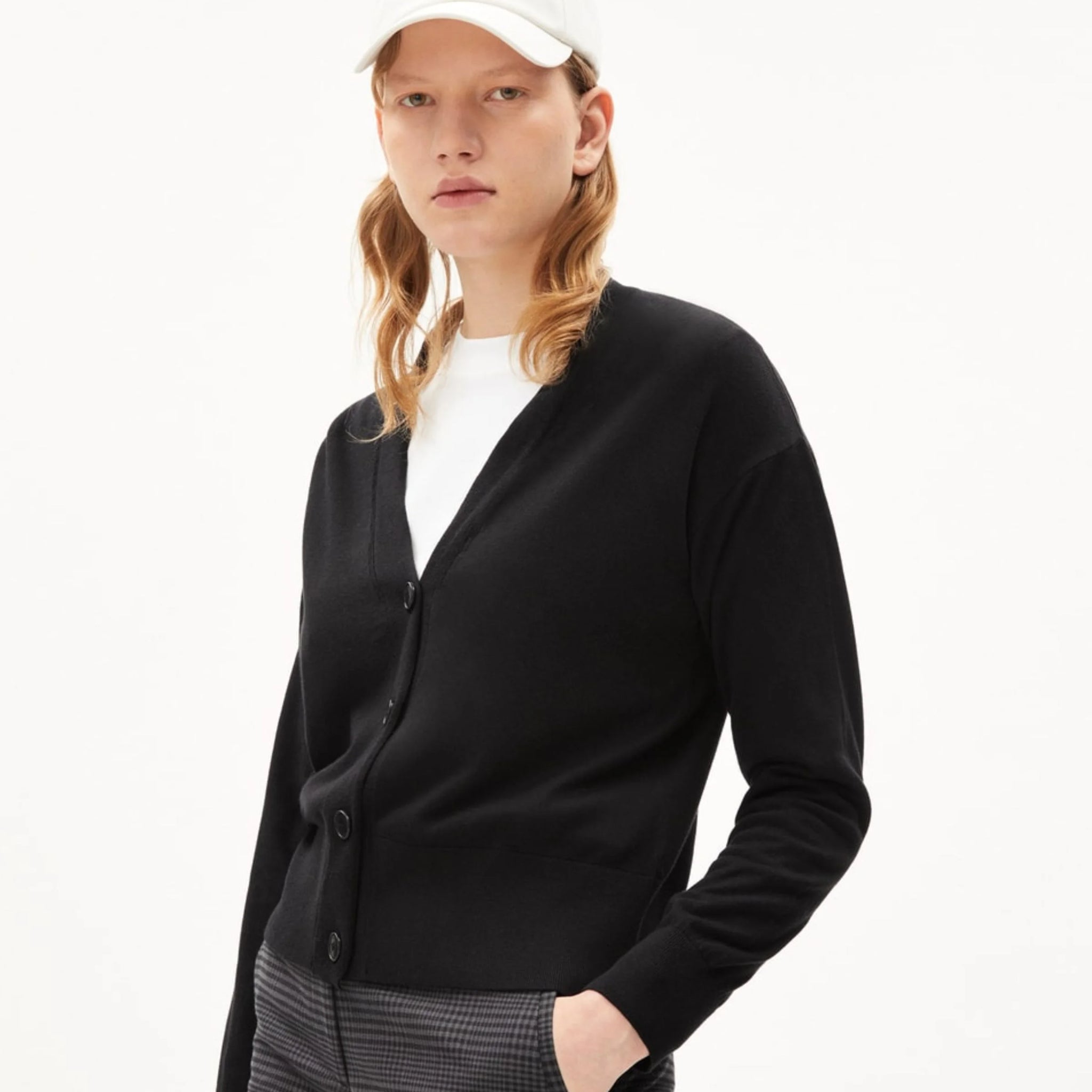 Person mit schwarzer Strickjacke, weißem Hemd und Basecap vor weißem Hintergrund.